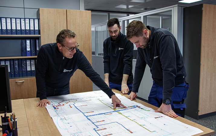 Drei Mitarbeiter von Kratschmayer stehen an einem Tisch und besprechen eine technische Zeichnung.